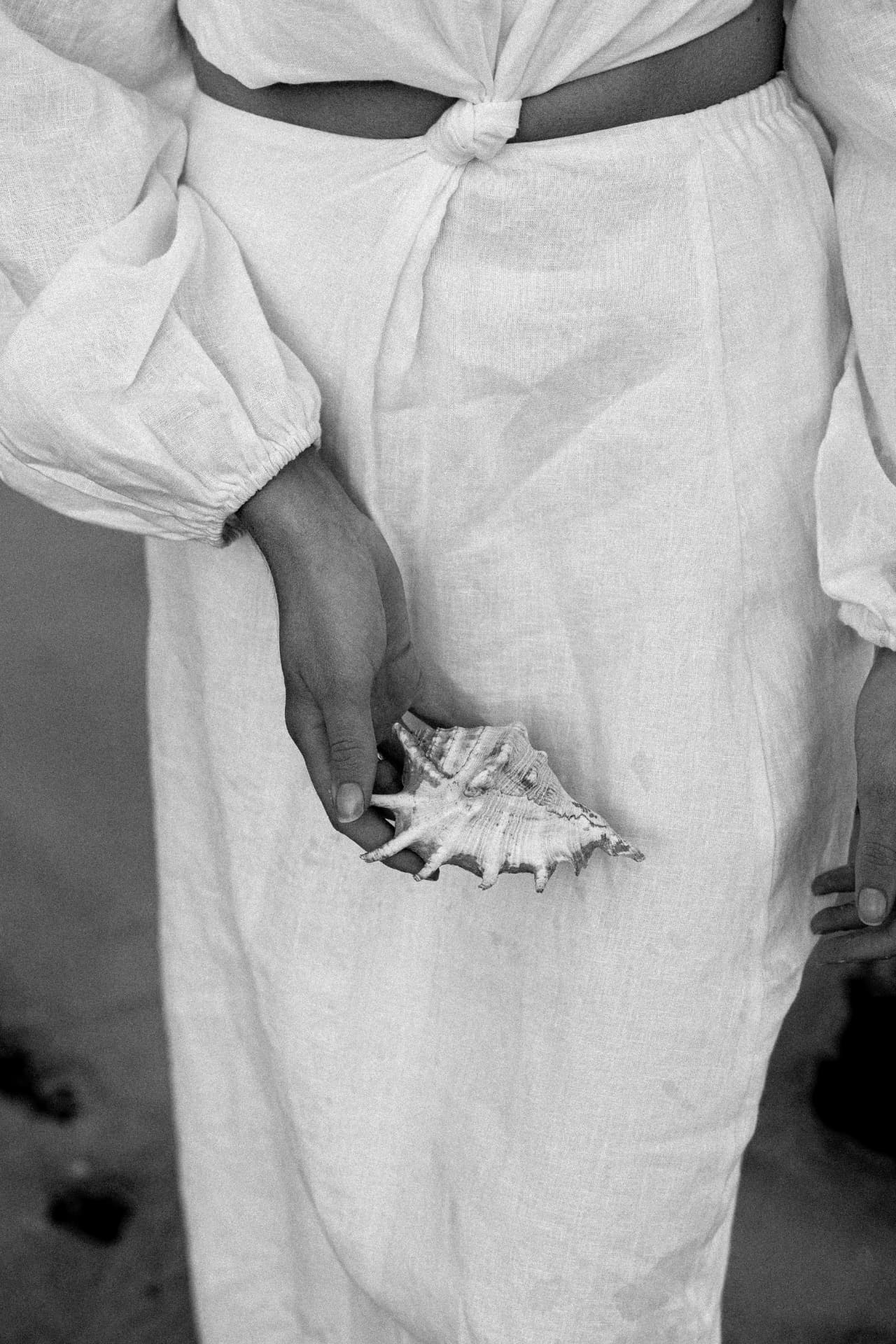 A woman in a white linen dress holding a shell at dusk